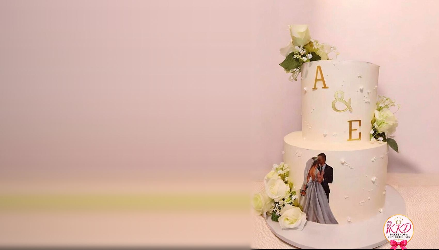 A two tier white wedding cake with gold monogram letters and fresh white roses, made by KKD Bakeshop