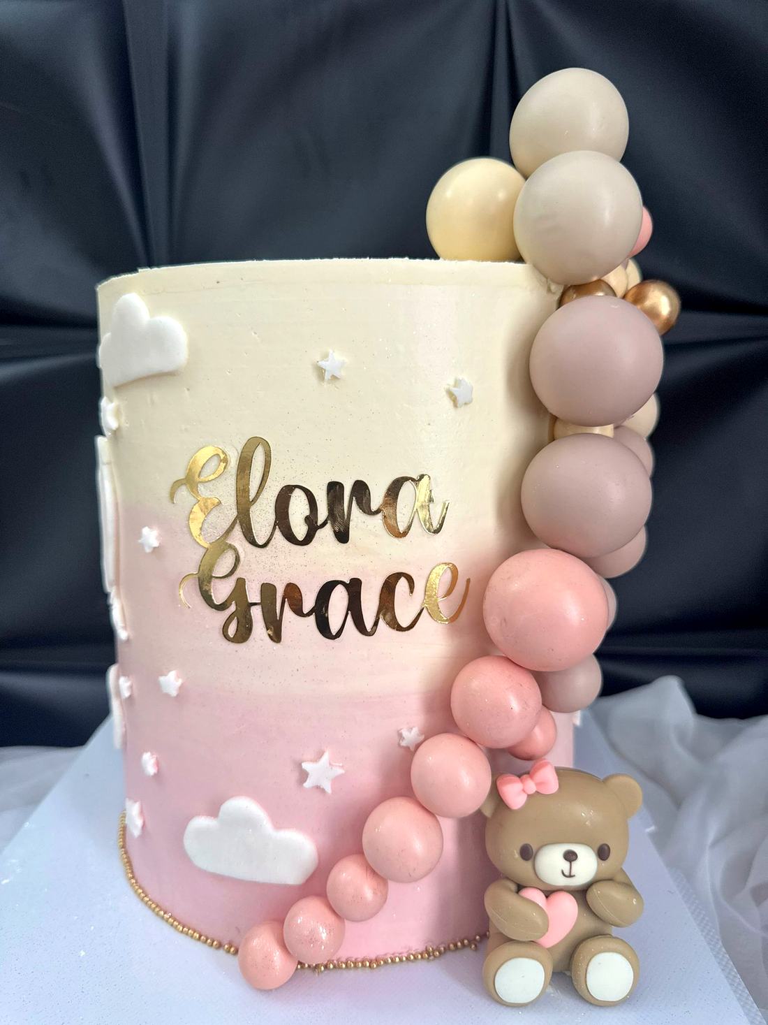A pink ombre celebration cake with a balloon garland and a teddy topper