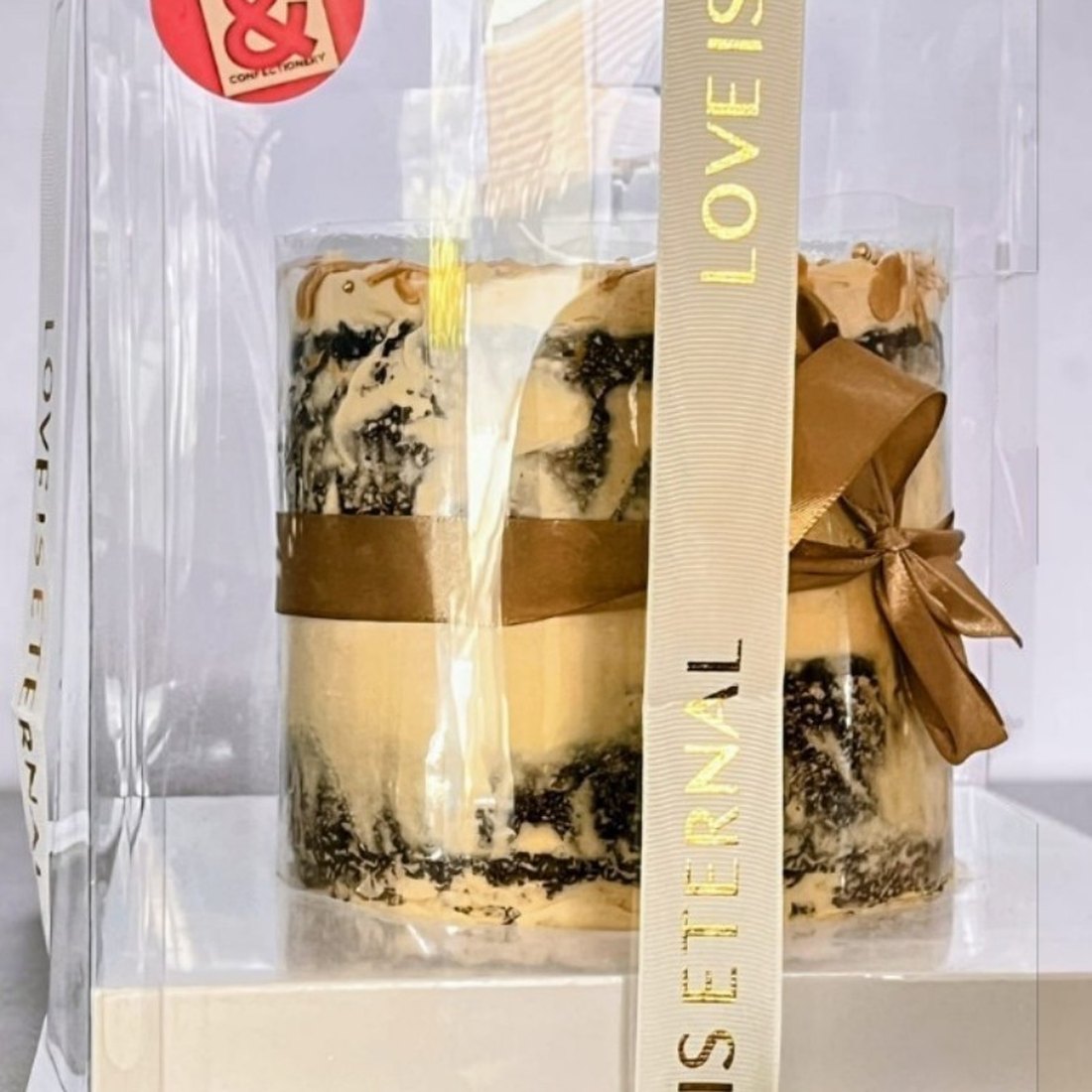 The cake in its signature packaging: an acetate collar inside a clear box, finished with a caramel ribbon