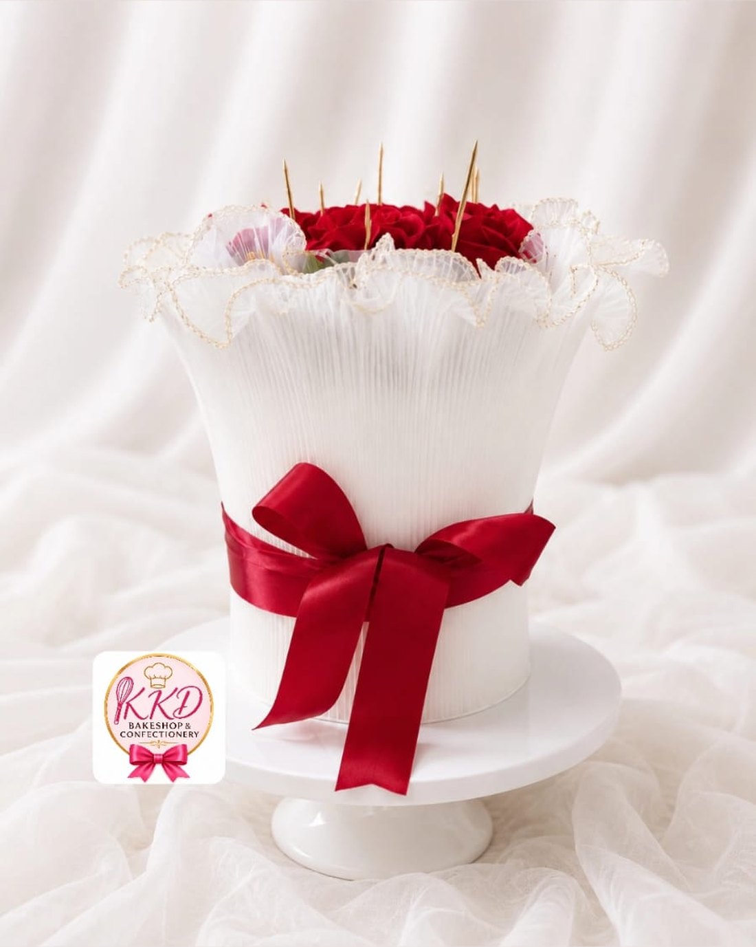 A tall white cake with a pleated finish, a gold-edged frill collar, a red satin bow and red roses on top